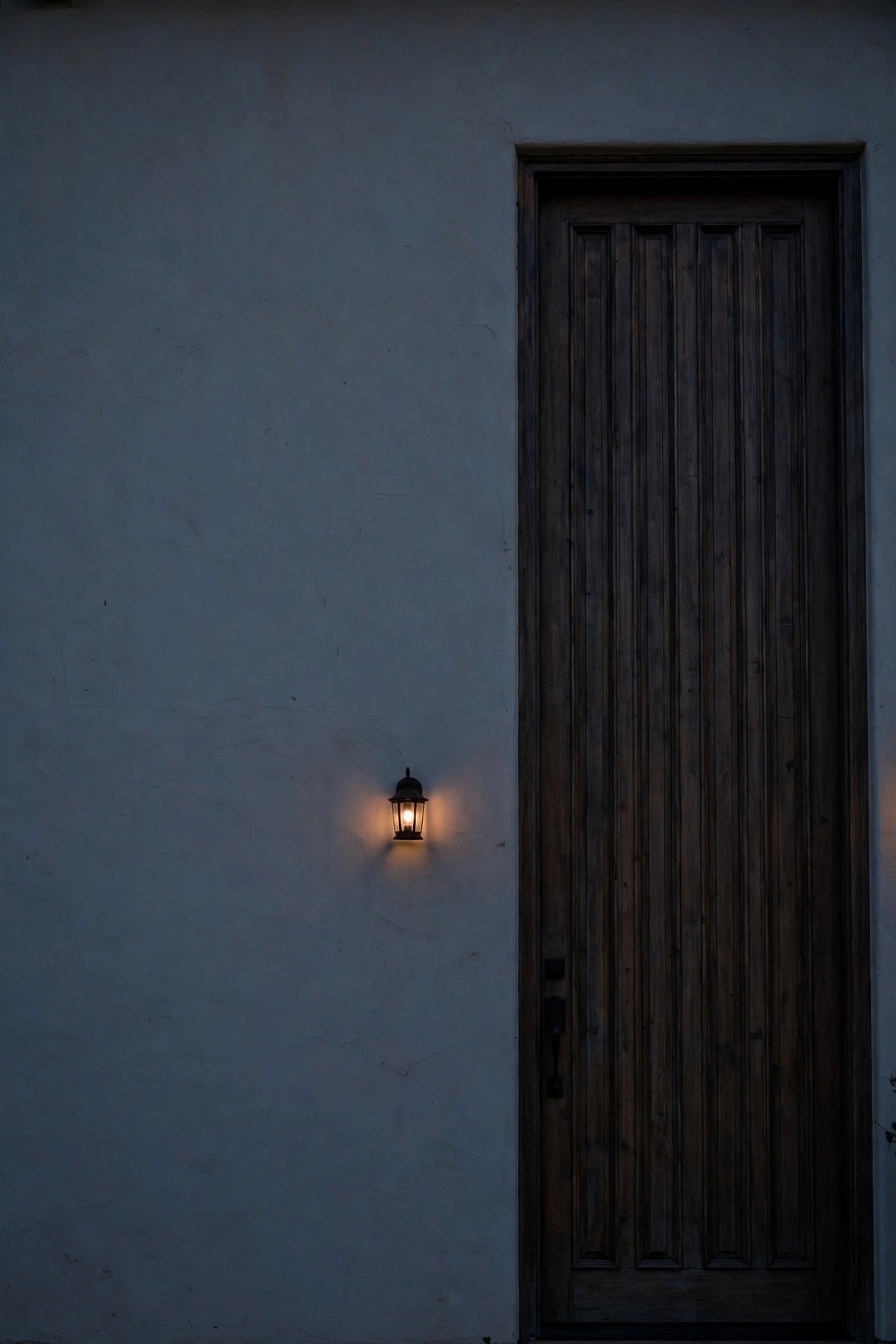 Small single wall lantern dwarfed beside a tall front door at dusk, its warm glow too weak for the large blank entry wall around it