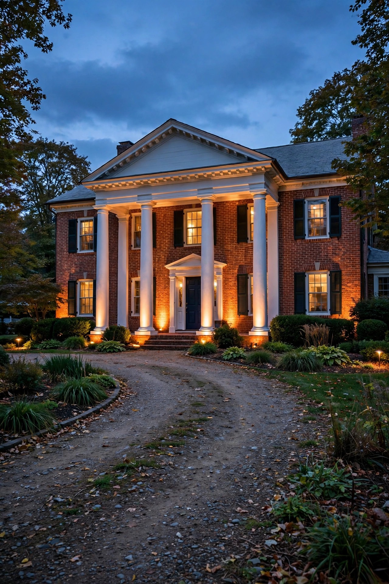Warm uplights washing the brick facade and white columns of a colonial home viewed from the driveway at dusk