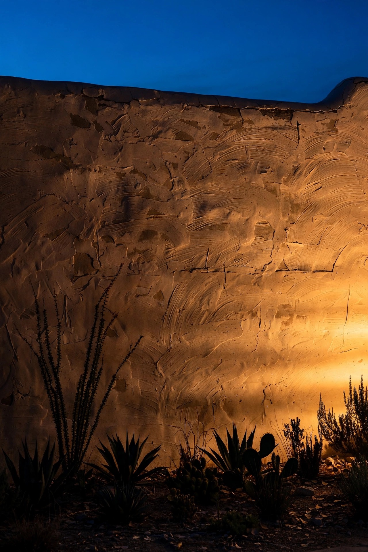 Adobe garden wall at night washed in warm grazing light that reveals hand-troweled texture, desert plants silhouetted at its base